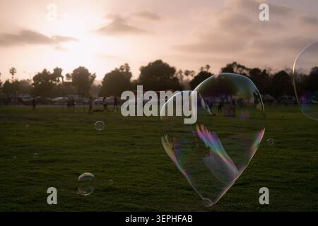 Large soap bubbles floating over a grassy field at sunset with people and trees in the background, creating a calm outdoor summer scene. Stock Photo