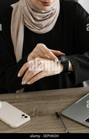 A woman wearing a hijab and black top interacts with her smartwatch displaying a heart rate reading while seated at a wooden desk with a laptop and ph Stock Photo