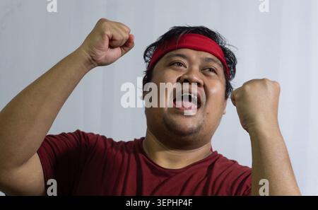 Indonesian male sports supporter cheering enthusiastically with both fists raised high in the air, wearing a red shirt and red headband standing again Stock Photo