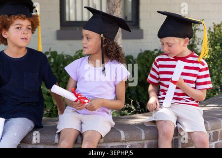 Diverse children sitting on curved brick bench at school wearing caps with tassels holding diplomas Stock Photo