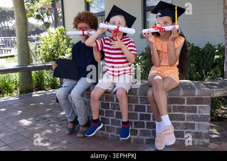 Diverse children sitting on brick ledge in schoolyard, wearing mortarboard caps, holding diplomas Stock Photo