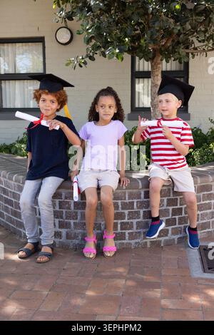 Three children sitting on low brick planter in courtyard wearing caps and holding red diplomas Stock Photo