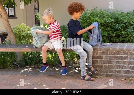 Two school-age boys sitting on wooden bench in shaded courtyard, sorting backpacks and chatting Stock Photo
