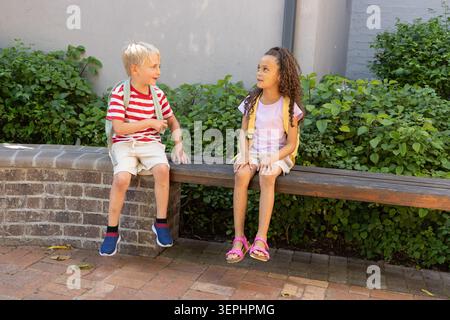 Two children sitting on wooden bench atop brick planter in sunny courtyard with backpacks Stock Photo