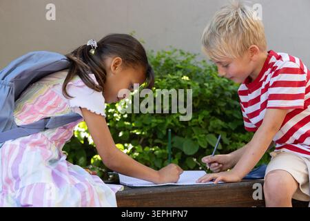 Two children sitting on wooden bench at schoolyard, writing in open notebook with pencils Stock Photo