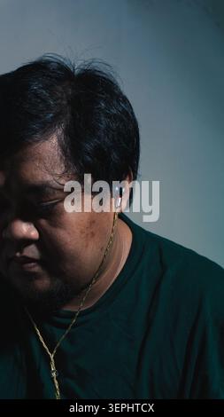An Indonesian adult male in a dark green shirt looks down with closed eyes while wearing wired earphones to show a calm and reflective mood against a Stock Photo