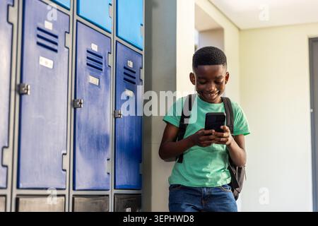 African American school-age boy leaning on column, using smartphone by blue lockers, copy space Stock Photo