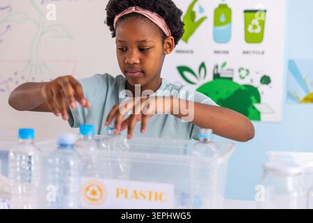 African American girl wearing tee pink-headband sorting bottles blue caps into PLASTIC bin at table Stock Photo