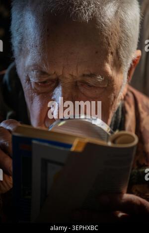An elderly man reads with scrutiny the text in a book,  using a magnifying glass Stock Photo