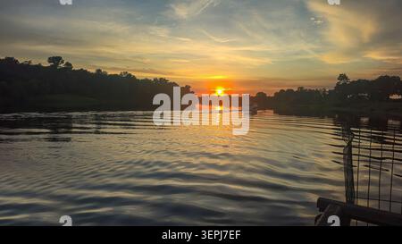 A beautiful summer sunset over a calm river. The orange and gold hues of the sky are reflected in the water's surface, and the silhouettes of the tree Stock Photo