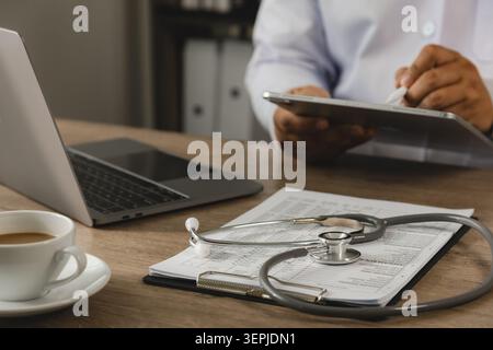 Healthcare professional reviewing patient information on digital tablet in a modern medical office with laptop, stethoscope, and clinical documents. C Stock Photo