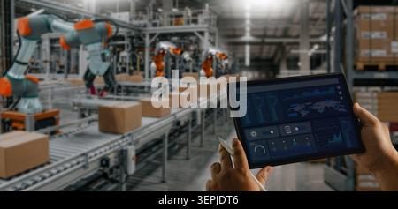 Operator monitoring automated warehouse operations on a tablet with robotic systems and conveyor lines handling packages, representing smart warehouse Stock Photo