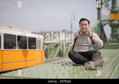 Young Asian man with headphones sits on a city bridge Stock Photo