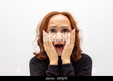 Sixty year old Asian woman looking surprised with hands on face. Mature lady expressing shock and amazement. Female portrait demonstrating strong emot Stock Photo