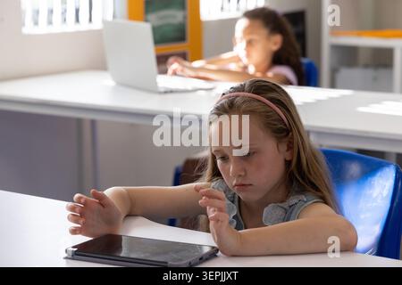 Female elementary students sitting at school desks front girl wearing pink headband using tablet Stock Photo