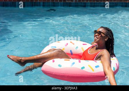 Adult African woman reclining on pink donut float, wearing red swimsuit and sunglasses in pool. Water, poolside, inflatable, sunny, vibrant, shades, locs Stock Photo