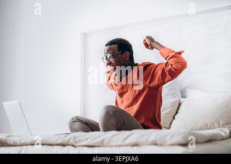 Joyful African-American guy in casual clothes and glasses puts hands up sitting near open laptop on comfortable bed at home Stock Photo