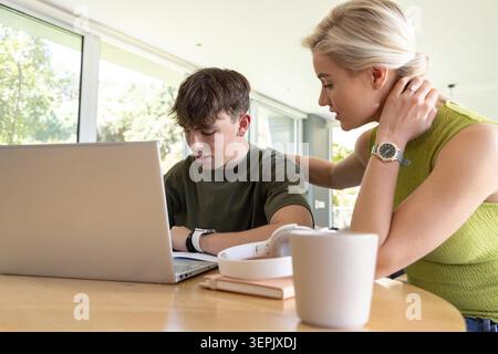Mother and teen son sitting at wooden dining table, using silver laptop and writing in notebook. Study, tutoring, guidance, parent, minimalist, daylight, window Stock Photo
