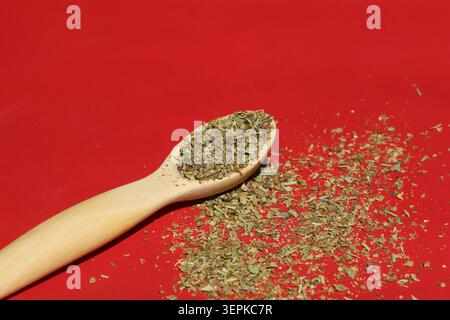 Angled closeup of dried crushed oregano herbs on a wooden spoon with scattered flakes over a solid red background Stock Photo