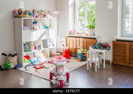 Messy kids bedroom interior featuring a large cubby bookshelf loaded with stuffed animals, books, and scattered educational toys on a rug Stock Photo