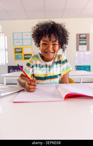 African American child in striped tee holding red pencil at school desk, smiling over red notebook Stock Photo