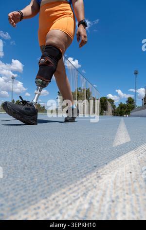 Woman amputee athlete walking on a blue running track with a prosthetic leg, focusing on training and determination under clear sky Stock Photo
