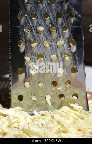 Front view closeup of pale yellow cheese shreds piled on a wooden surface below a stainless steel metal grater Stock Photo