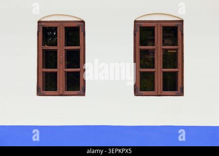 Symmetrical minimal facade with pair of brown wooden windows on white wall with blue base stripe Stock Photo