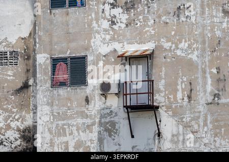 A close-up view of an old, weathered building wall featuring peeling paint, a small window with a cloth inside, an air conditioning unit, and a rustic Stock Photo