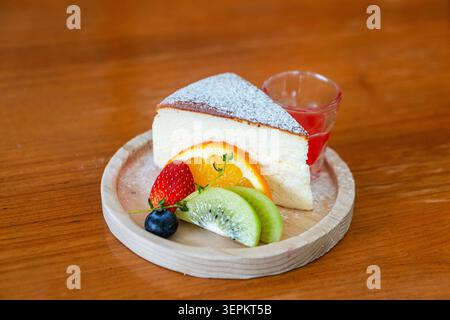 A delicate slice of cake served on a wooden plate alongside fresh fruit and a small glass of red jelly, perfect for breakfast or dessert enjoyment. Stock Photo