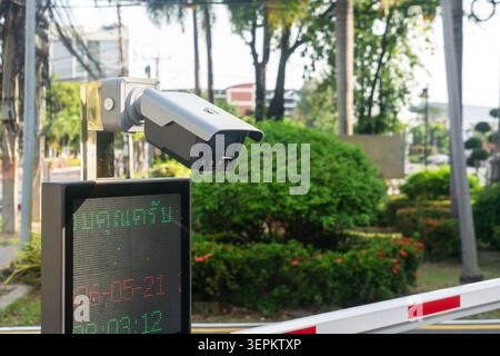This image features a security surveillance camera mounted outdoors in a park-like setting with trees, bushes, and urban elements, providing safety an Stock Photo