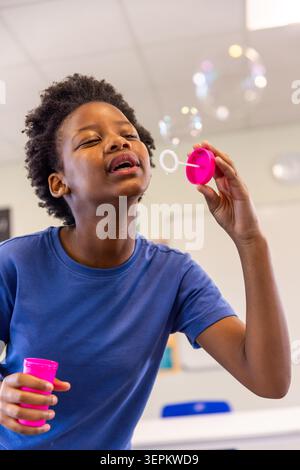 African American boy standing in classroom blowing soap bubbles with pink wand and container Stock Photo
