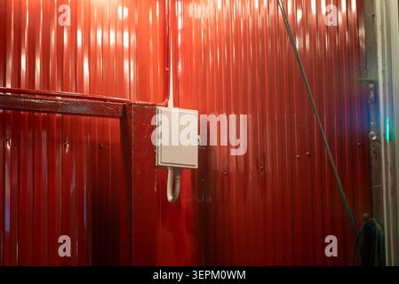 This image features a red corrugated metal wall indicating an industrial or construction setting, with an electrical box mounted and a hose extending Stock Photo