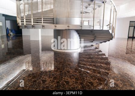 This image shows a modern interior space featuring a spiral staircase with metal railings, standing on a highly polished, reflective marble floor that Stock Photo