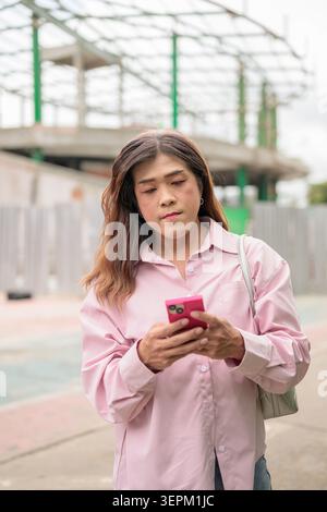 A vertical medium shot of a mid adult transgender Thai businesswoman from the Isan region looking down at her pink smartphone while overseeing an urba Stock Photo