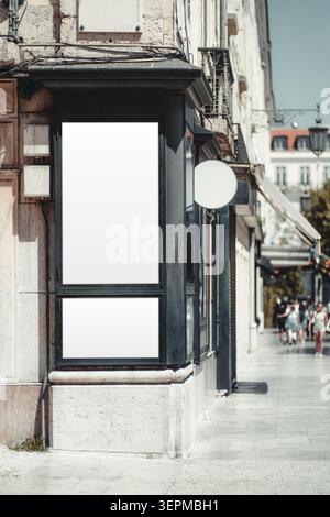 Empty rectangular poster template on a black storefront facade with round projecting sign, old city street, sidewalk and blurred pedestrians in sunny Stock Photo