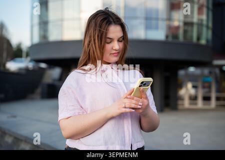 Young businesswoman using a smartphone outside a modern office building in an urban workplace setting. Digital communication and mobile technology use Stock Photo