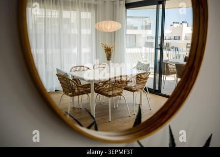 A bright modern dining area reflected in a large round mirror with a wooden frame, showing a white dining table surrounded by rattan chairs, Spain Stock Photo