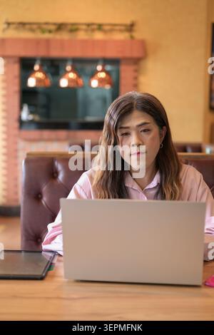 A vertical medium shot of a mid-adult Thai transgender businesswoman from the Isan region looking at her laptop screen while working remotely at a res Stock Photo