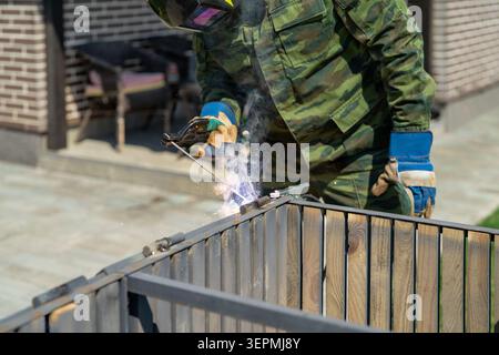 Man in protective welding mask operating welding machine in outdoor residential yard. Performing repair of storage box, home maintenance, safety equip Stock Photo