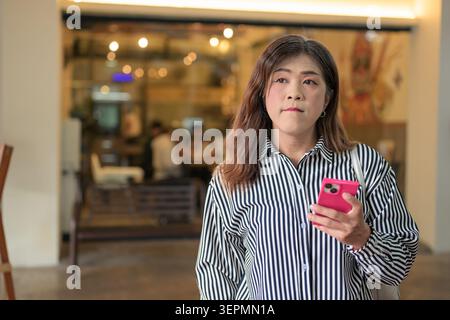 A horizontal medium shot of a mid-adult Thai Isan transgender businesswoman looking off-camera while holding her pink smartphone in a restaurant entry Stock Photo