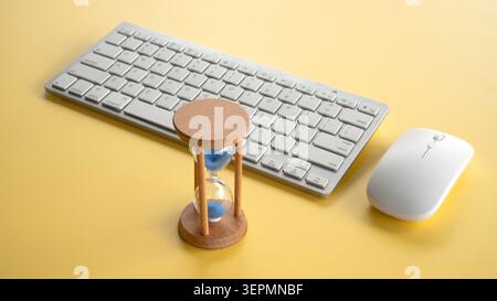 Hourglass standing next to a modern computer keyboard and mouse on a bright yellow background, representing time and technology. Stock Photo
