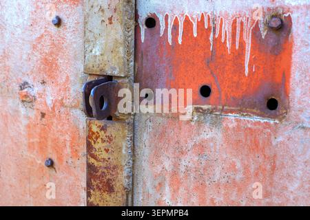 Rusty metal door latch and steel padlocking lug are welded on a garage gate. Weathered orange iron surface texture displays old paint drips, bolts, co Stock Photo