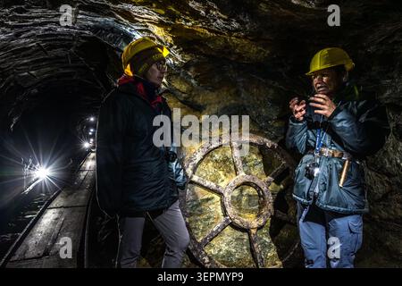 Two participants on the guided tour are chatting at the Hochfeld exhibition mine in front of a historic wheel from the mine’s former haulage system. In the background, the tracks of the mine railway lead deeper into the illuminated tunnel. Rosental, Neukirchen am Großvenediger, Bezirk Zell am See, Salzburg, 5741, Austria Stock Photo