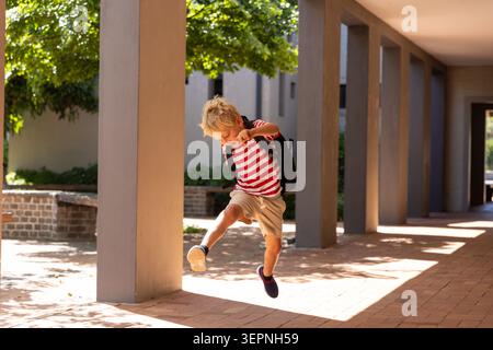 Boy child jumping beneath walkway with concrete columns in brick courtyard, wearing backpack Stock Photo