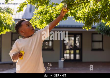 African American boy child blowing bubbles under branch holding yellow bottle, wand in courtyard Stock Photo