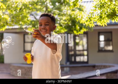 African American boy blowing soap bubbles under leafy tree at sunlit courtyard with yellow wand Stock Photo