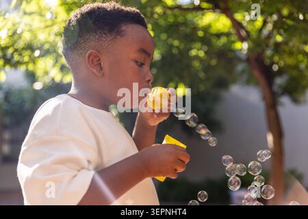 African American boy child wearing white shirt blowing bubbles with yellow wand and bottle in yard Stock Photo