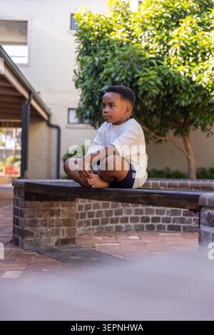 African American child wearing white T-shirt, sitting on brick bench in sunny courtyard, barefoot Stock Photo