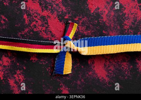 Ribbons in the colors of the German and Ukrainian flags, tied in a strong knot on a black and red textured background as a symbol of friendship, unity Stock Photo